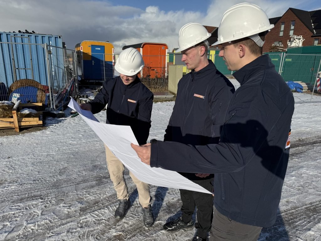 Drie vakmensen van Bouwsector Nederland met witte bouwhelm bestuderen een tekening op een nieuwbouwterrein